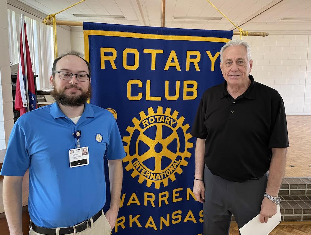 Two men standing next to a banner that says rotary club of warren arkansas.
