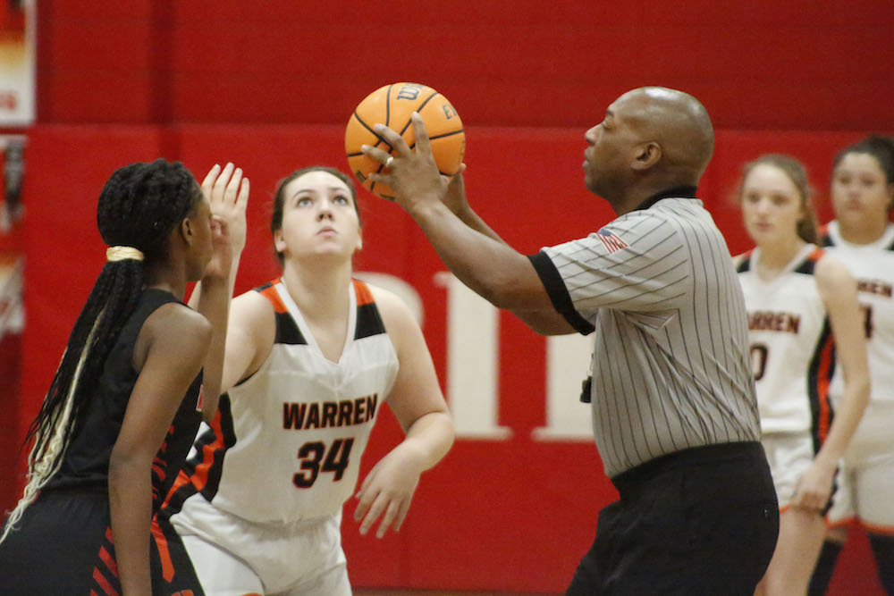 A referee holding a basketball over two players.