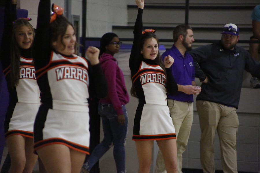 Two cheerleaders are standing in a gym with other people.