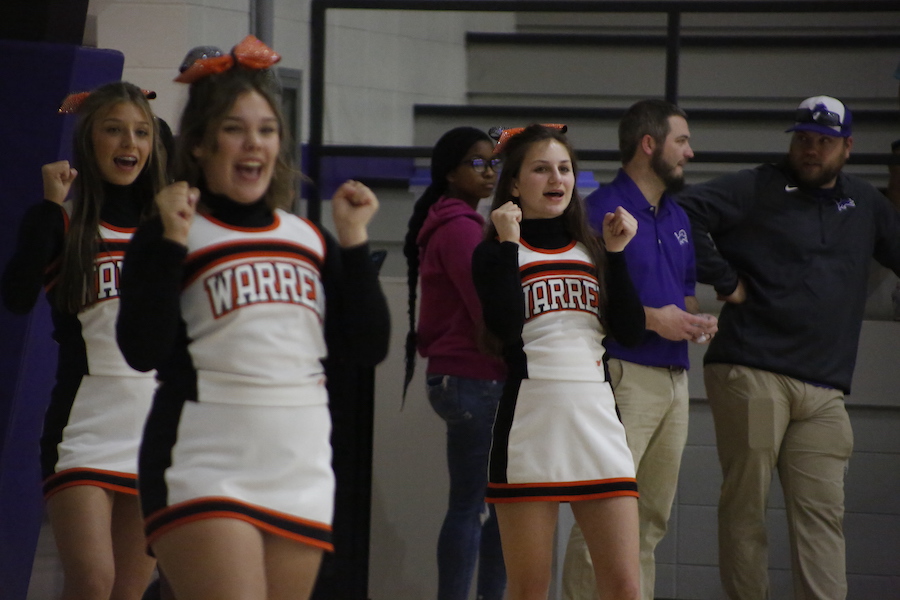 Two cheerleaders are standing in a gym with their hands raised.