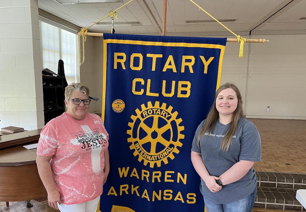 Two women standing next to a banner that says rotary club warren arkansas.