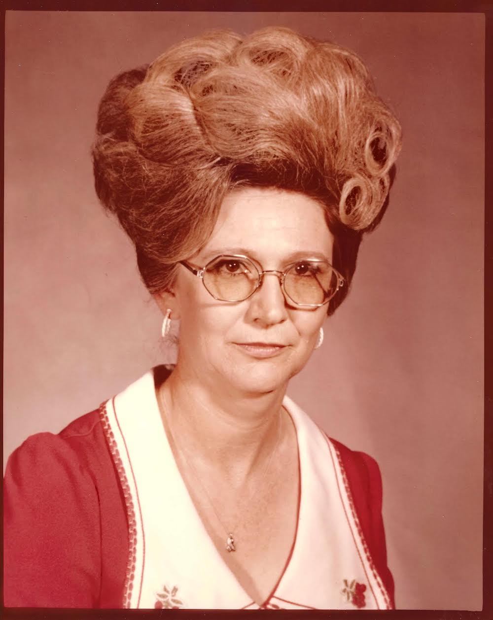A woman with big hair and glasses in front of a picture.