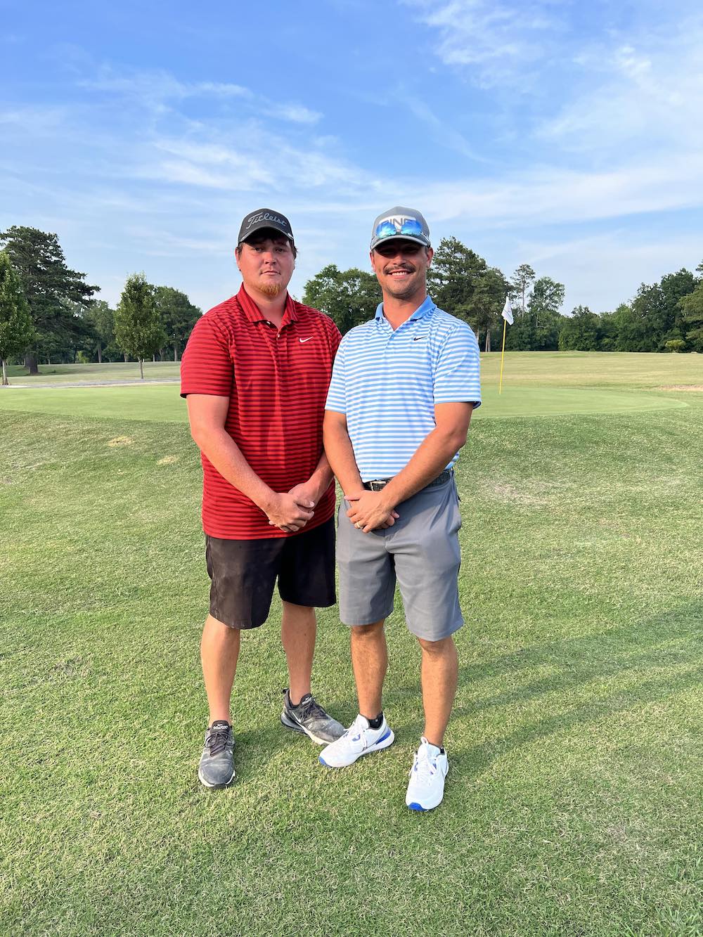 Two men standing on a golf course with trees in the background.