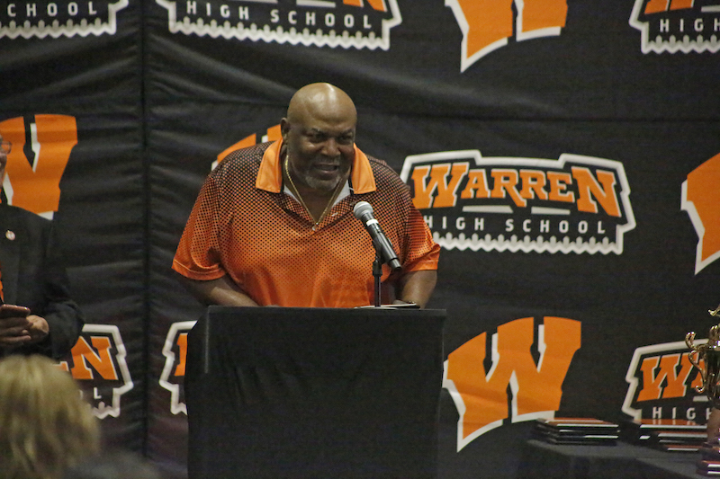 A man in an orange shirt is speaking at a podium.