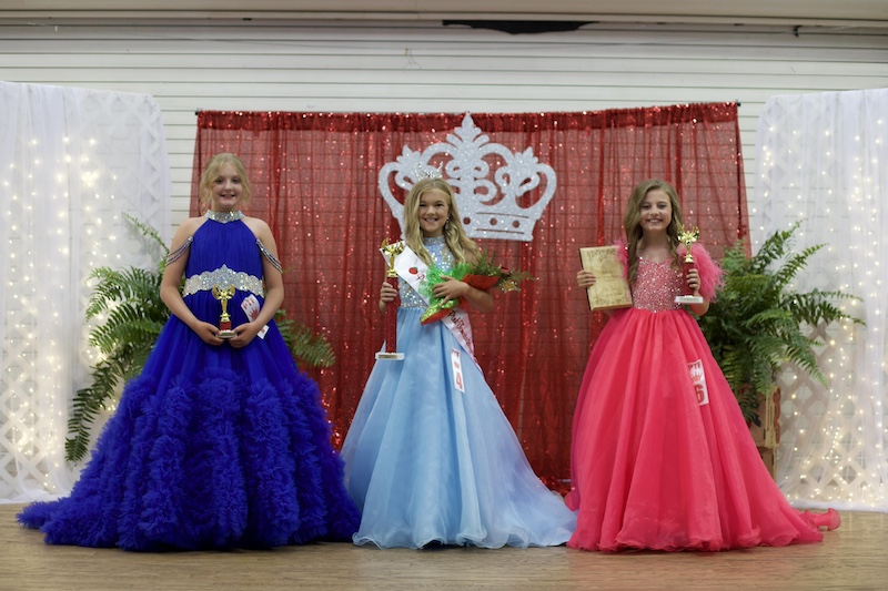 Three girls in dresses and a crown on the stage.