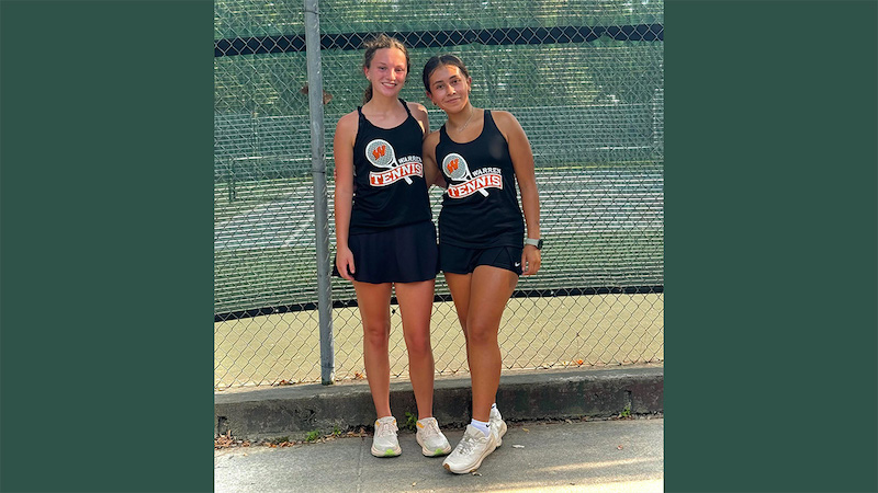 Two girls standing next to each other on a tennis court.