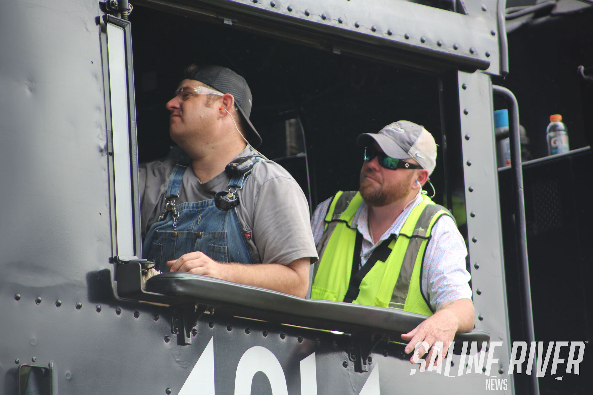 Two men in safety vests and hard hats look out a window.