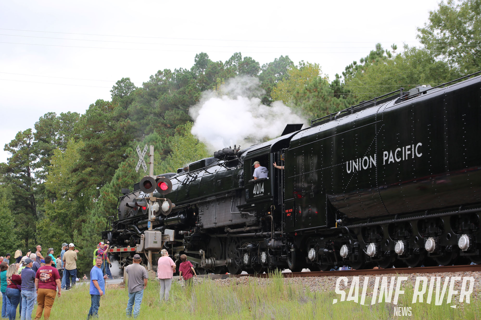 A train engine with passengers on it and people standing around.