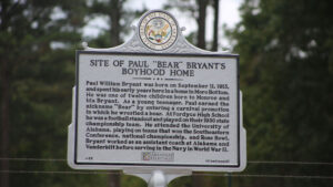 A sign in front of trees with a building behind it.