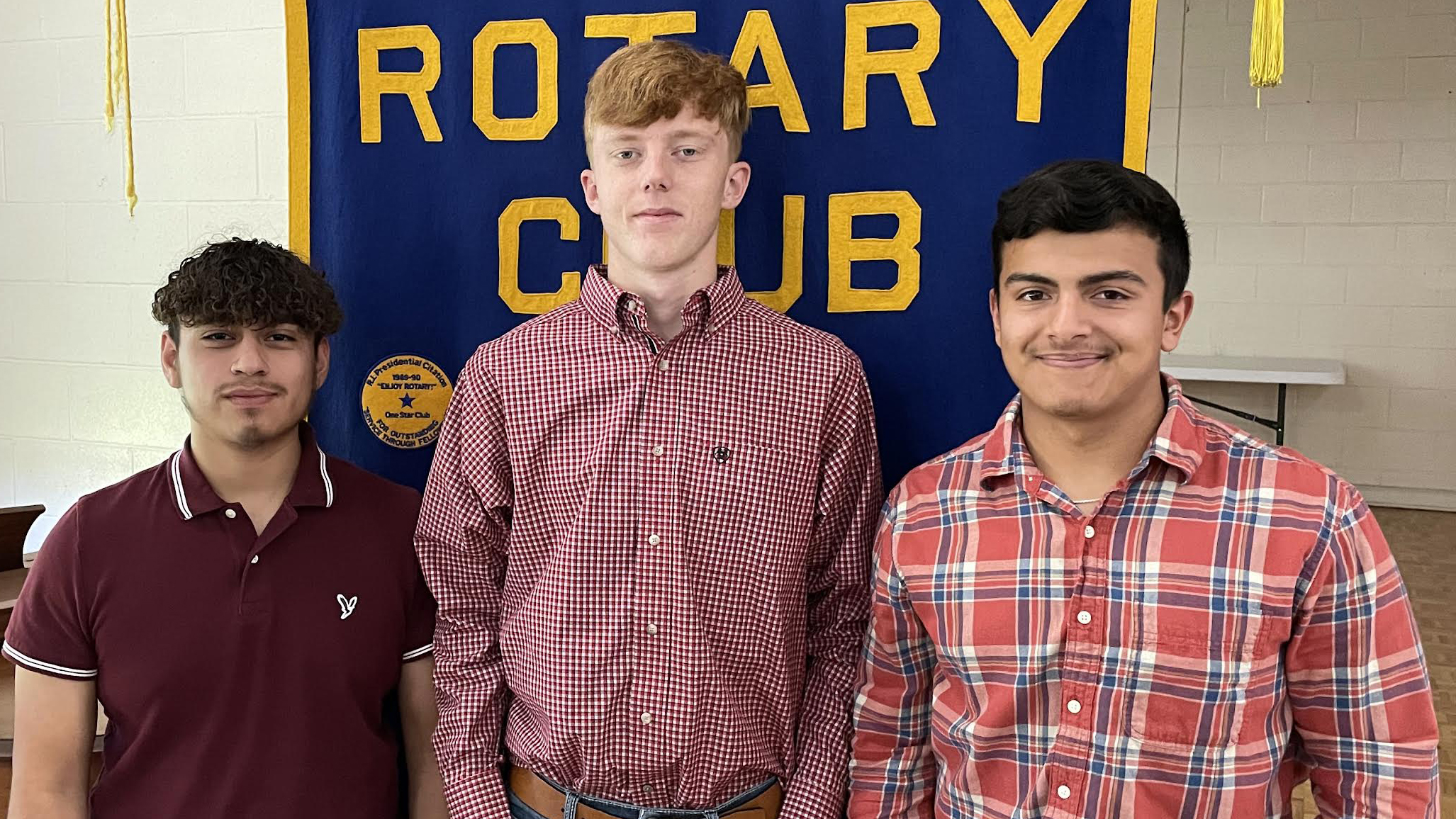Three young men standing in front of a rotary club banner.