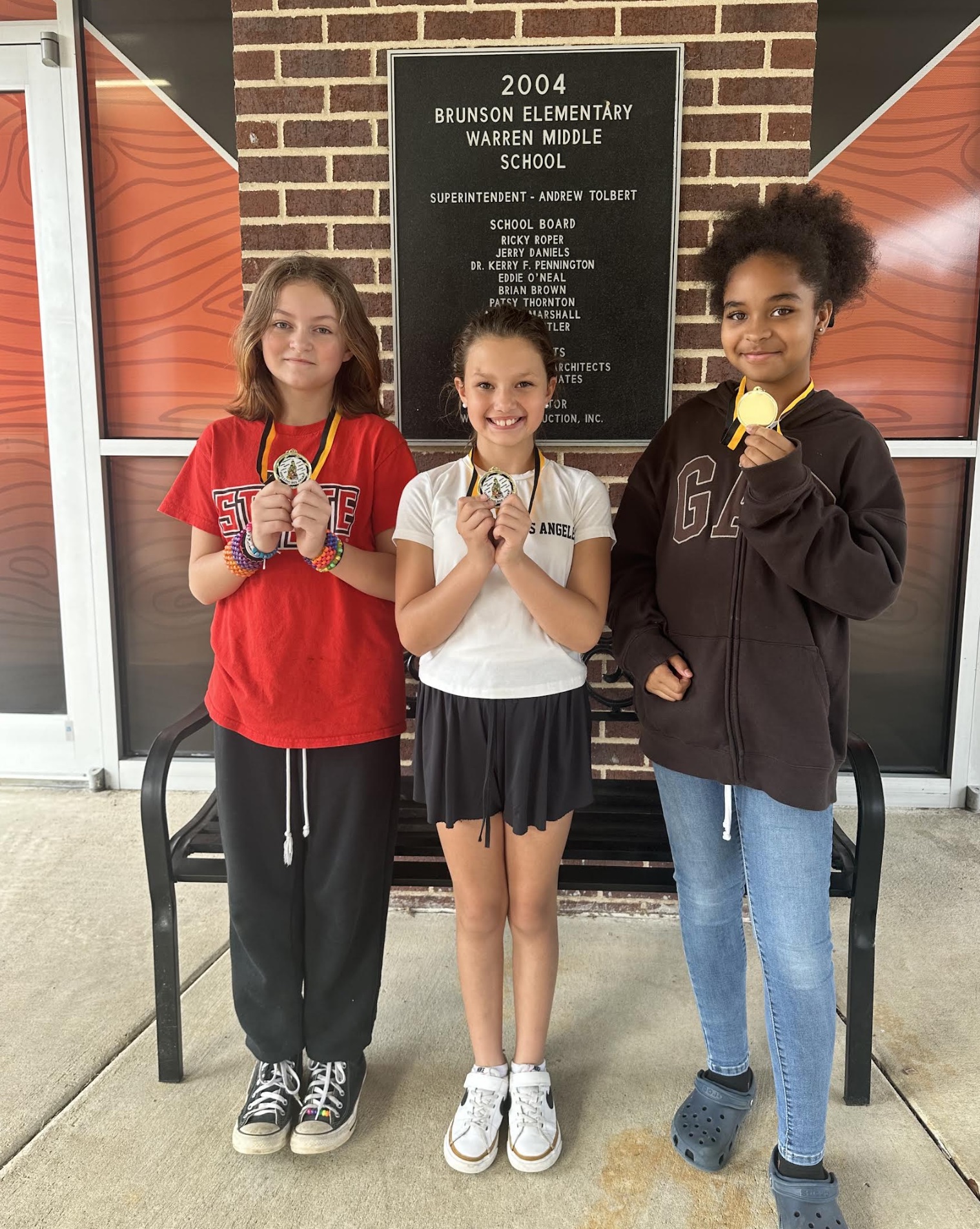 Three girls holding medals in front of a brick wall.