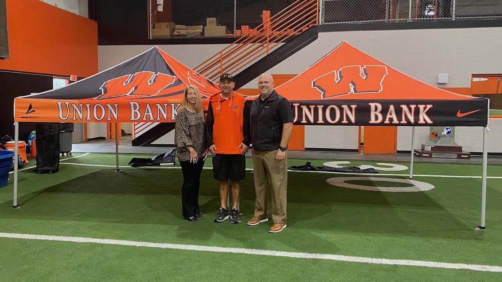 Three people standing on a field with an orange and black sign behind them.