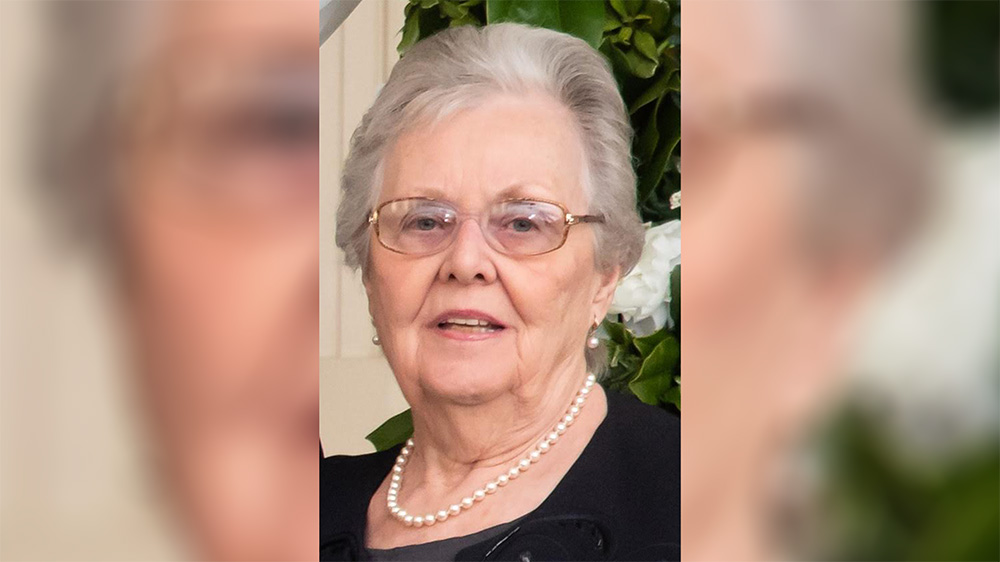 A woman with glasses and pearls in front of flowers.