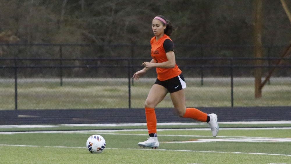 A girl in orange shirt kicking a soccer ball.