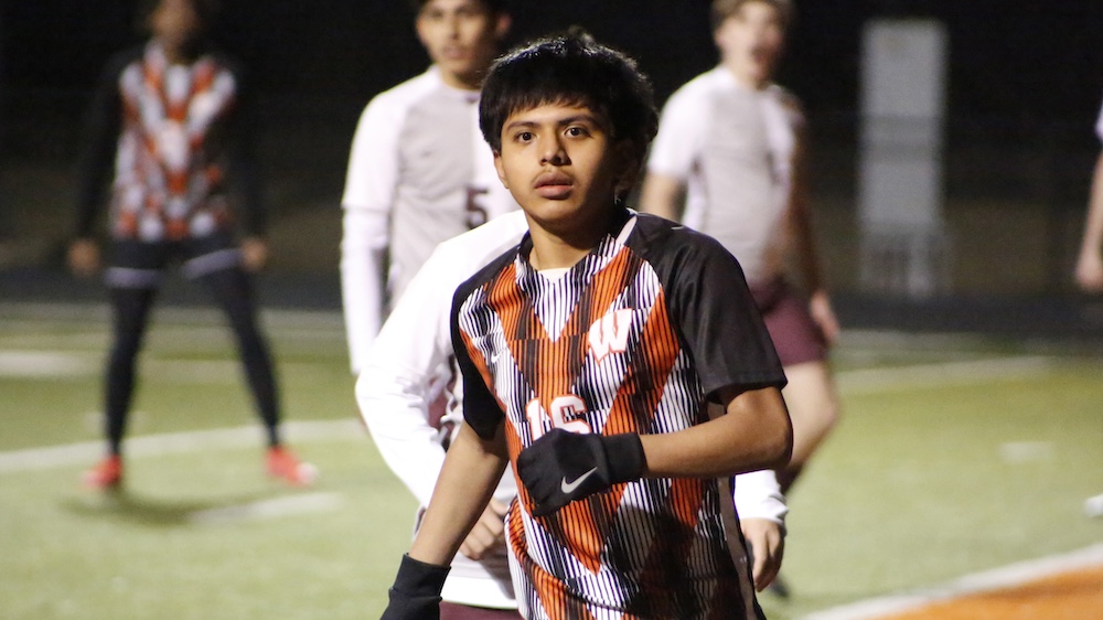 A young man in a soccer uniform stands on the field.